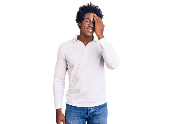 Hombre afroamericano guapo con cabello afro usando ropa casual y gafas que cubren un ojo con la mano, sonrisa confiada en la cara y emoción sorpresa.  - Foto, imagen