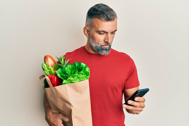 Красивий чоловік середнього віку тримає мішок продовольства, використовуючи смартфон, розслаблений серйозним виразом обличчя. простий і природний погляд на камеру
.  - Фото, зображення
