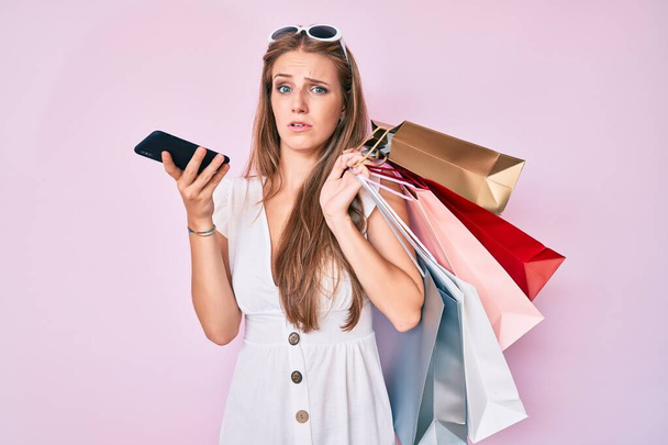 Молодая блондинка держит сумки с покупками разговаривая на смартфоне невежественное и запутанное выражение лица. Концепция сомнений.  - Фото, изображение