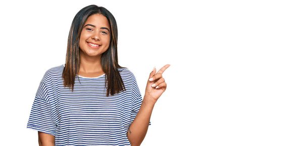 Νεαρό Λατίνο κορίτσι που φοράει καθημερινά ρούχα με ένα μεγάλο χαμόγελο στο πρόσωπο, δείχνοντας με το χέρι και το δάχτυλο στο πλάι κοιτάζοντας την κάμερα.  - Φωτογραφία, εικόνα
