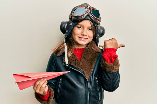 Kleines schönes Mädchen in Pilotenuniform mit Papierflieger, der den Daumen zur Seite zeigt und glücklich mit offenem Mund lächelt  - Foto, Bild