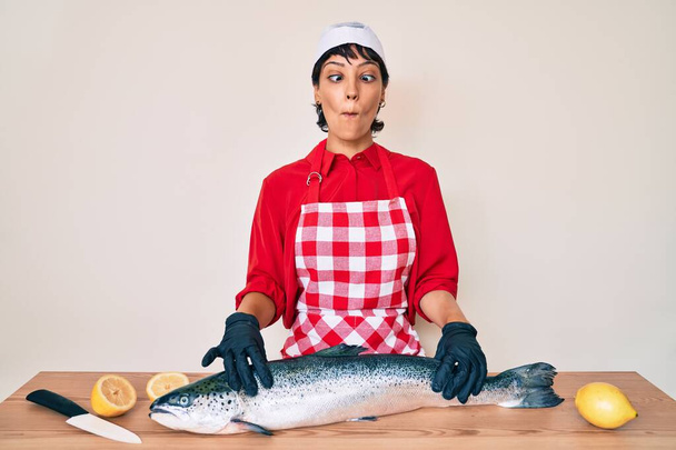 Hermosa mujer morena pescadero cocinar salmón crudo fresco haciendo cara de pescado con la boca y los ojos entrecerrados, loco y cómico.  - Foto, Imagen