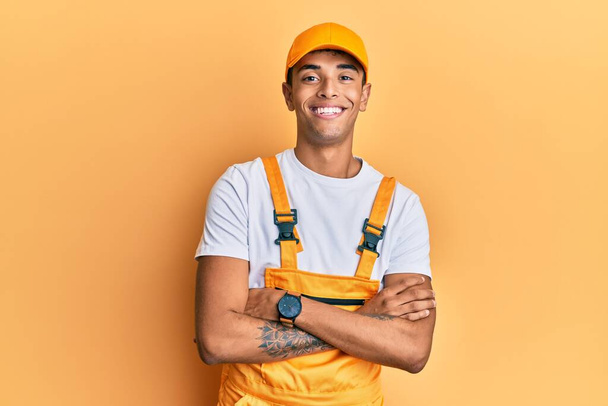 Jeune bel homme afro-américain portant l'uniforme de bricoleur sur fond jaune heureux visage souriant avec les bras croisés regardant la caméra. personne positive.  - Photo, image