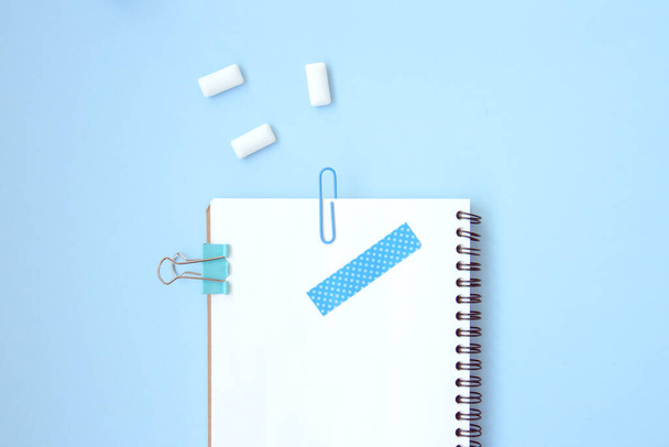 White open notebook with blue paper clip, clip and piece of scotch tape and gum on blue background. Blue set stationery with space for text. School stationery. Flat lay - Photo, Image