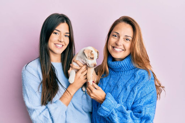Όμορφη ισπανόφωνη μητέρα και κόρη χαμογελώντας χαρούμενη αγκαλιά μικρό σκυλί πάνω από απομονωμένο ροζ φόντο. - Φωτογραφία, εικόνα