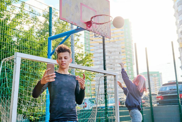 Guy teenager recording video stream using smartphone, on sports street court, girl playing basketball - Foto, Imagen