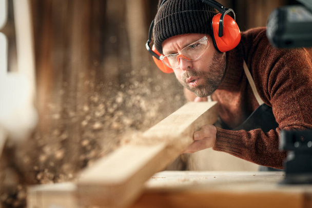 Adult male woodworker in protective goggles and headphones blowing sawdust from wooden detail while working in carpentry workshop - Фото, изображение