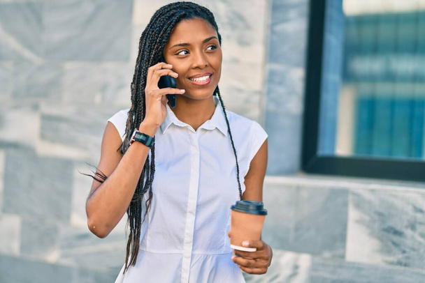 Молода афроамериканська жінка, яка розмовляє на смартфоні і п "є, забирає каву в місто.. - Фото, зображення