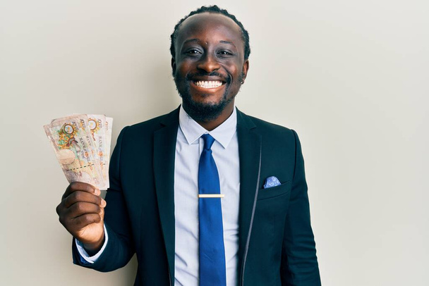 Handsome young black man wearing business suit holding pounds banknotes looking positive and happy standing and smiling with a confident smile showing teeth  - 写真・画像