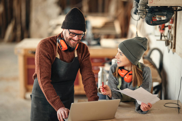 Positive man and woman in aprons and with protective headphones standing near table with laptop and discussing ideas for project while working together in professional carpentry workshop - Photo, Image