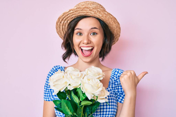 Mladá krásná dívka v letním klobouku drží květiny ukazující palec nahoru na stranu s úsměvem šťastný s otevřenými ústy  - Fotografie, Obrázek