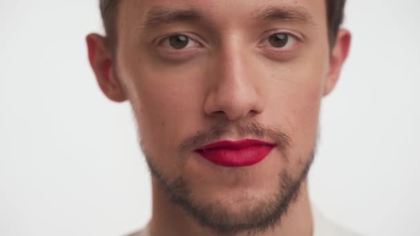 Portrait of glamorous man with beard, painted lips with bright red lipstick. Close-up of bearded guy smiles, winks with brown eyes, flirts, looks straight ahead like in a mirror on white background. - Footage, Video