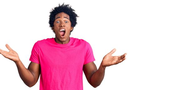 Bonito homem americano africano com cabelo afro vestindo camiseta rosa casual celebrando a vitória com sorriso feliz e expressão vencedora com as mãos levantadas  - Foto, Imagem