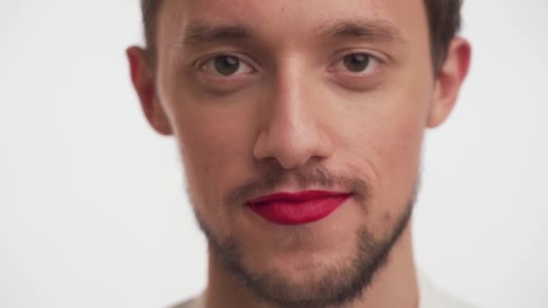 Jonge Spaanse man met donkerbruine baard, snor, lippen geschilderd in rode vrouwenlippenstift loopt rond, beweegt zijn ogen in een cirkel, aan verschillende kanten, glimlachend op witte achtergrond van dichtbij.. - Video
