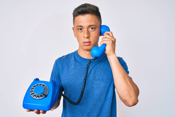 Νεαρός Ισπανόφωνος κρατώντας το παλιό τηλέφωνο χαλαρό με σοβαρή έκφραση στο πρόσωπο. απλή και φυσική κοιτάζοντας την κάμερα.  - Φωτογραφία, εικόνα