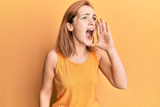 Junge kaukasische Frau in lässigem Stil mit ärmellosem Hemd, die schreit und laut schreit, mit der Hand auf dem Mund. Kommunikationskonzept.  - Foto, Bild