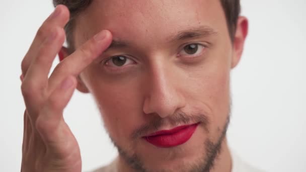 Όμορφος μετροσέξουαλ ή γκέι άντρας με γενειάδα, φοράει κόκκινο κραγιόν στα χείλη, κοιτάζει την κάμερα με καστανά μάτια, χαϊδεύει το μέτωπό του με δάχτυλο σε λευκό φόντο. Κοντινό πορτραίτο ενός γοητευτικού αρσενικού. - Πλάνα, βίντεο