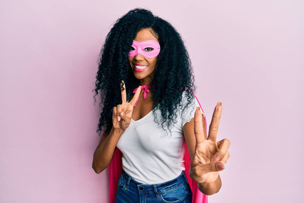 Középkorú afro-amerikai nő szuperhős jelmezt visel, mosolyog, és a kamerába néz, amint ujjakat mutat győzelmi jelekkel. Második..  - Fotó, kép