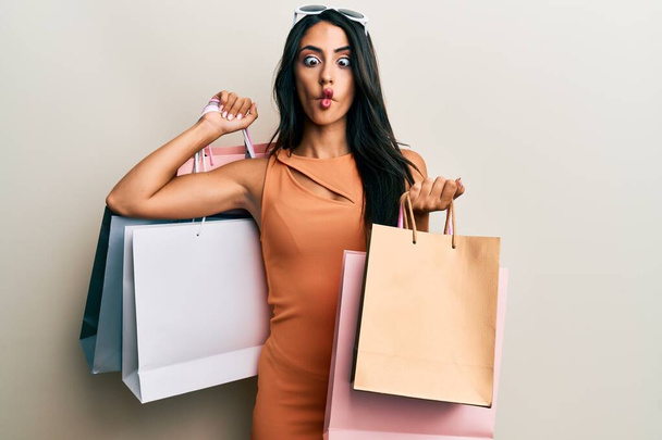 Beautiful hispanic woman holding shopping bags making fish face with mouth and squinting eyes, crazy and comical.  - Фото, зображення