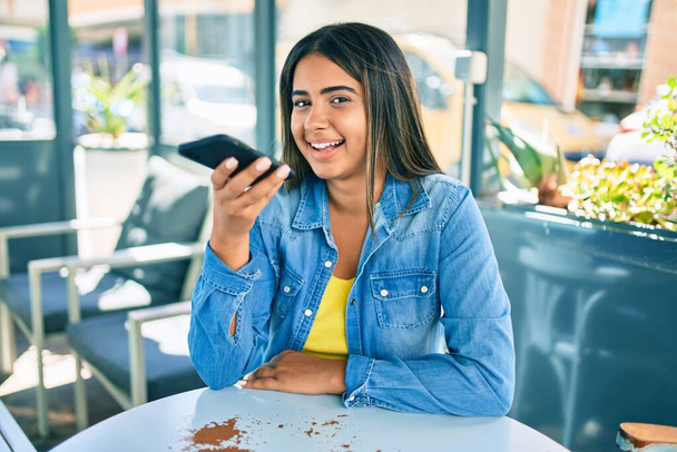 Młoda Latynoska uśmiechnięta szczęśliwa wysyłając wiadomość dźwiękową za pomocą smartfona na tarasie kawiarni. - Zdjęcie, obraz