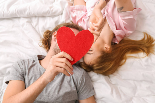 Fiatal pár, vörös szívvel az ágyon fekve, felülnézetből. Valentin-napi ünneplés - Fotó, kép