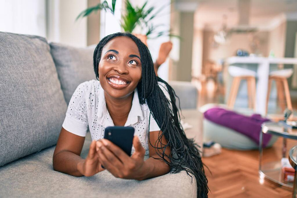 Fiatal afro-amerikai nő mosolyog boldog segítségével smartphone feküdt a kanapén otthon - Fotó, kép