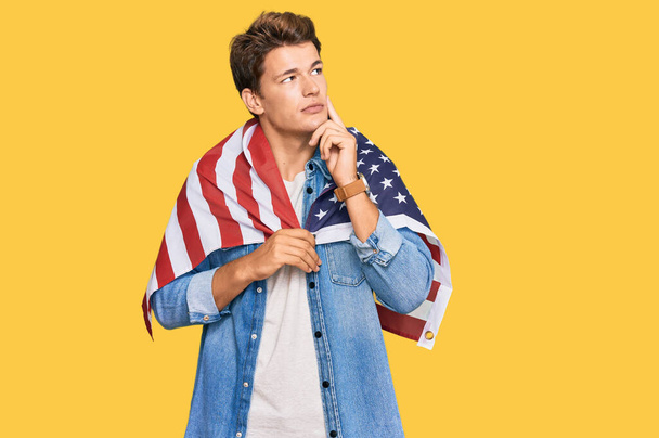 Handsome caucasian man holding united states flag serious face thinking about question with hand on chin, thoughtful about confusing idea  - Foto, Imagem