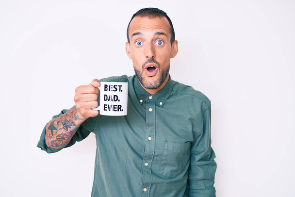 Молодий красивий чоловік з татуюванням п'є чашку кави з найкращим татом коли-небудь повідомленням наляканий і вражений відкритим ротом для несподіванки, обличчя віри
  - Фото, зображення
