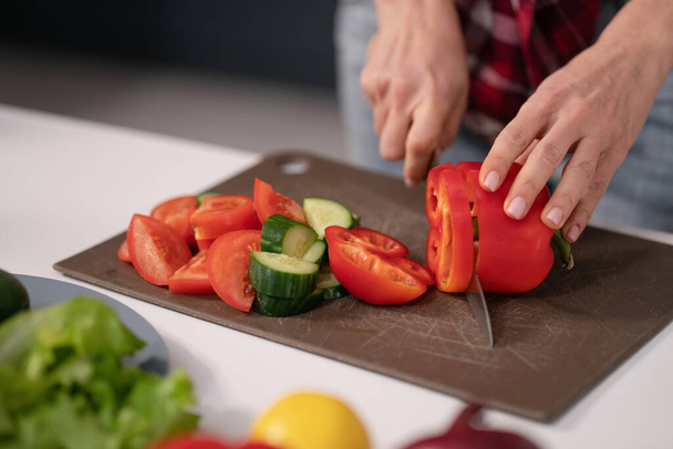 Cuts vegetables for a fresh salad young housewife cut bell pepper, cucumber, tomato on cutting board preparing for a family dinner standing in the new kitchen of a new home. Healthy lifestyle - 写真・画像