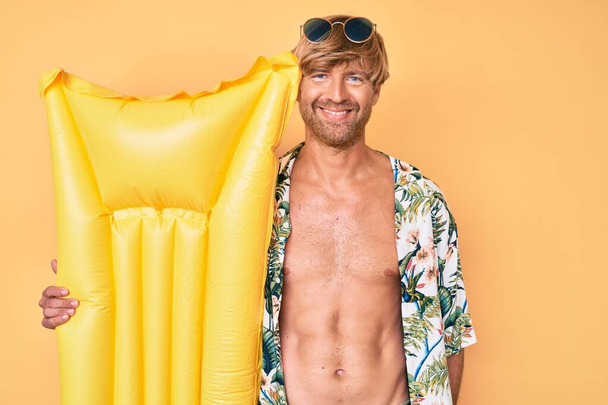 Mladý blonďatý muž v letním stylu a držení matrace float vypadá pozitivně a šťastně stojící a usmívá se s sebevědomým úsměvem ukazující zuby  - Fotografie, Obrázek