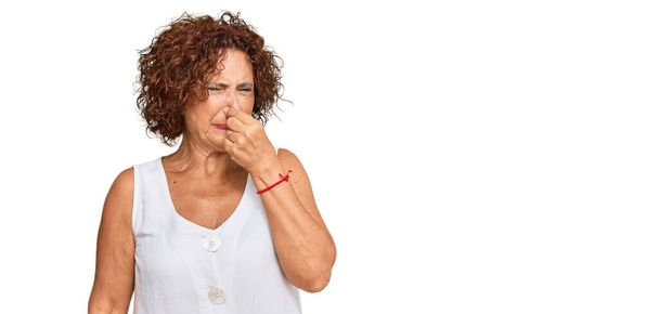 Beautiful middle age mature woman wearing casual white shirt smelling something stinky and disgusting, intolerable smell, holding breath with fingers on nose. bad smell  - Photo, Image