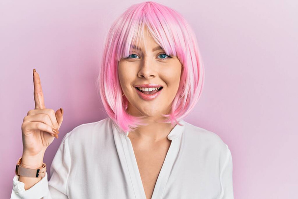 Junge blonde Frau mit pinkfarbener Perücke zeigt mit erhobenem Zeigefinger auf erfolgreiche Idee. aufgeregt und glücklich. Nummer eins.  - Foto, Bild