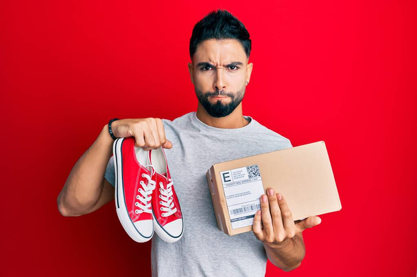 Young man with beard taking casual red shoes from box skeptic and nervous, frowning upset because of problem. negative person.  - Photo, Image