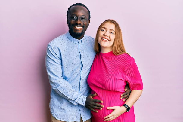 赤ん坊を期待している若い異人種間のカップルは、幸せとクールな笑顔で妊娠中の腹に触れています。運のいい人.  - 写真・画像