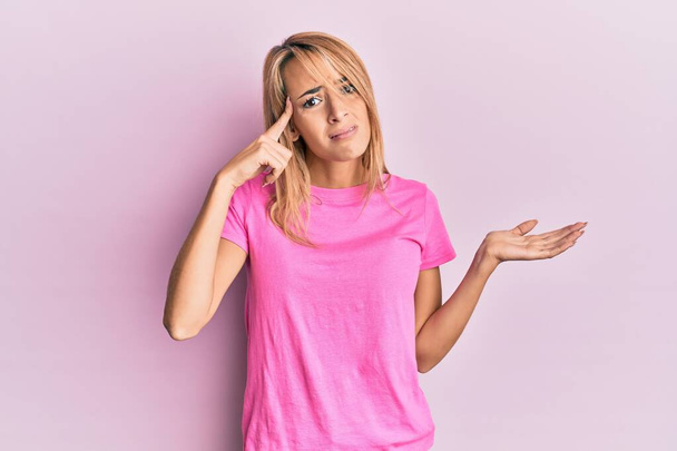 Beautiful blonde woman wearing casual pink tshirt confused and annoyed with open palm showing copy space and pointing finger to forehead. think about it.  - Photo, Image