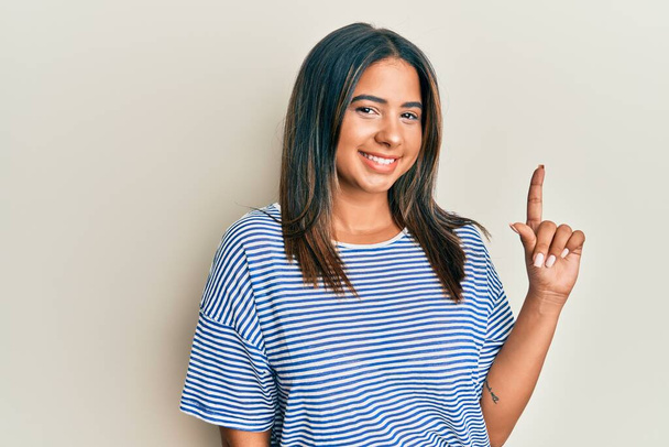 Молода латинська дівчина в повсякденному одязі посміхається з ідеєю або питанням, вказуючи пальцем вгору з щасливим обличчям, номер один
  - Фото, зображення
