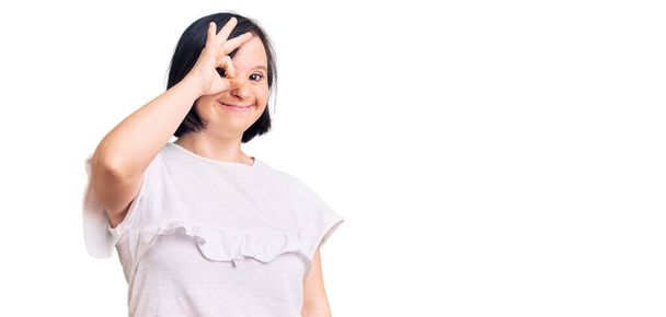 Brunette vrouw met down syndroom dragen casual wit tshirt doet ok gebaar met de hand glimlachen, oog kijken door vingers met gelukkig gezicht.  - Foto, afbeelding