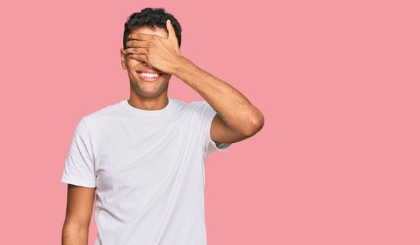 Jonge knappe Afro-Amerikaanse man draagt casual witte tshirt glimlachend en lachend met de hand op het gezicht bedekt ogen voor verrassing. blind concept.  - Foto, afbeelding