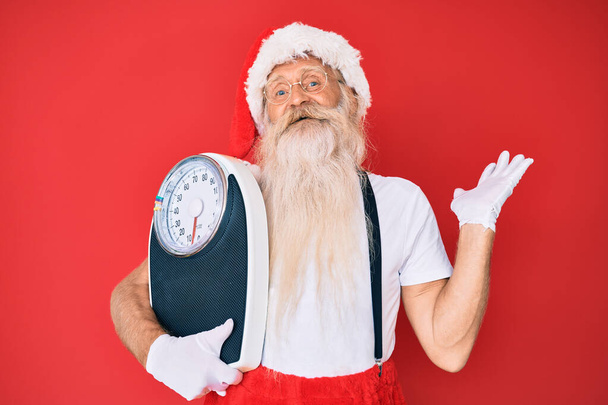 Старик на диете в традиционном костюме Санта-Клауса празднует победу со счастливой улыбкой и выражением победителя с поднятыми руками  - Фото, изображение
