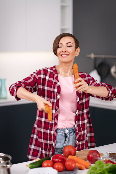 Throws up washed carrots trying to juggle and does not catch any charming short hair housewife prepares carrot cake in the kitchen. Healthy food at home. Healthy food leaving concept - Foto, afbeelding