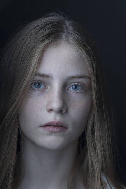 Charming blond young girl with freckles and blue eyes on a black background, indoors, close up portrait - Zdjęcie, obraz