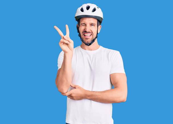 Jonge knappe man met een fietshelm lachend met een vrolijk gezicht knipogend naar de camera die het overwinningsbord doet. nummer twee.  - Foto, afbeelding
