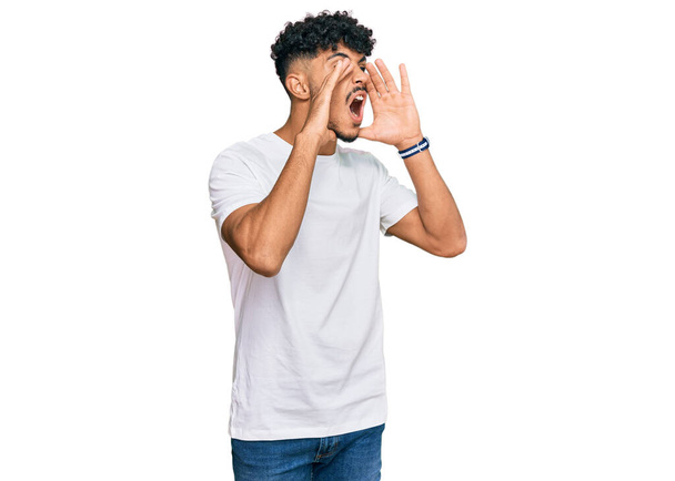 Jeune homme arabe portant casual t-shirt blanc criant en colère à haute voix avec les mains sur la bouche  - Photo, image