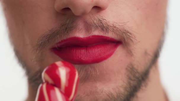 Close up mouth of gorgeous guy with make-up  red lipstick on lips. One young male with brown beard, moustache licking and sucking sweet sugar lollipop isolated on white background. Mouth view of man - Footage, Video