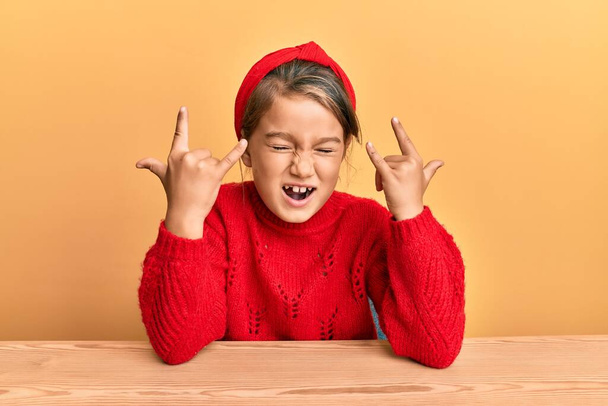 Little beautiful girl wearing casual clothes sitting on the table shouting with crazy expression doing rock symbol with hands up. music star. heavy concept.  - Фото, изображение