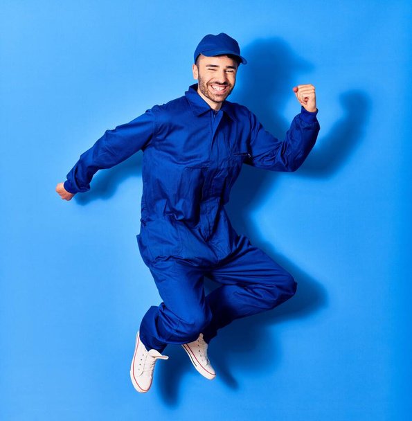 Young handsome hispanic man wearing painter uniform and cap smiling happy. Jumping with smile on face doig winner sign with fists up over isolated blue background - Photo, Image