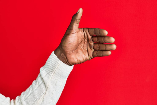 Arm und Hand eines schwarzen afrikanisch-amerikanischen jungen Mannes über rotem, isoliertem Hintergrund mit unsichtbarem Gegenstand, leere Hand beim Ausschneiden und Greifen  - Foto, Bild
