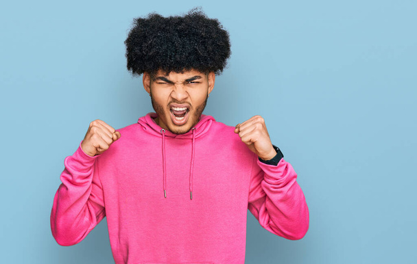 Młody, afrykański Amerykanin z afro włosami w luźnej różowej bluzie wściekły i wściekły podnoszący pięści sfrustrowany i wściekły krzycząc ze złości. wściekłość i agresywne pojęcie.  - Zdjęcie, obraz