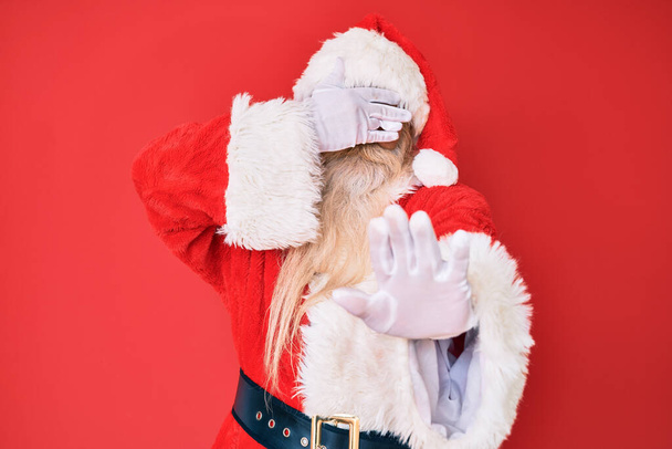 Alter älterer Mann mit grauen Haaren und langem Bart in traditionellem Weihnachtsmann-Kostüm, der die Augen mit Händen bedeckt und mit traurigem und ängstlichem Gesichtsausdruck Stopp-Gesten macht. Peinliches und negatives Konzept.  - Foto, Bild