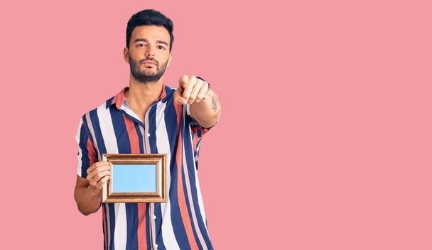 Jeune homme hispanique beau tenant le cadre vide pointant du doigt vers la caméra et vers vous, geste confiant et sérieux  - Photo, image